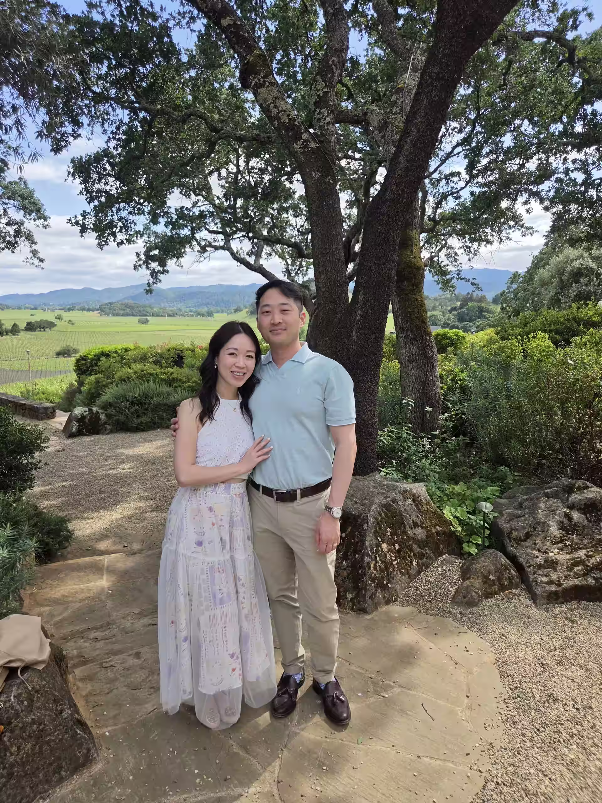 A couple standing beneath a large tree with vineyards and hills in the background.