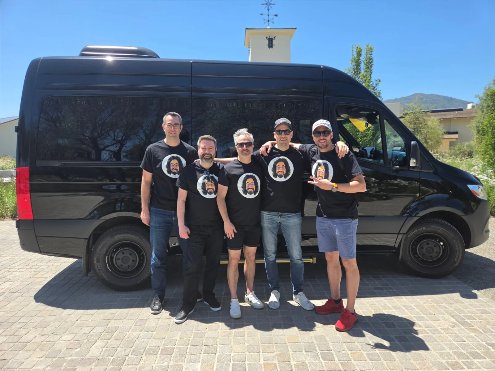 Five men beside a black Sprinter van in sunshine.